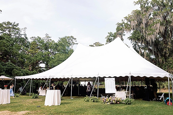 Wedding reception tent with string lights over cocktail tables and banquet seating on a lawn beneath oak trees draped in moss