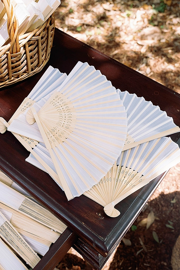 Wedding ceremony fans in a wicker basket on a wooden table, with paper hand fans wedding favors set in dappled sunlight outdoors