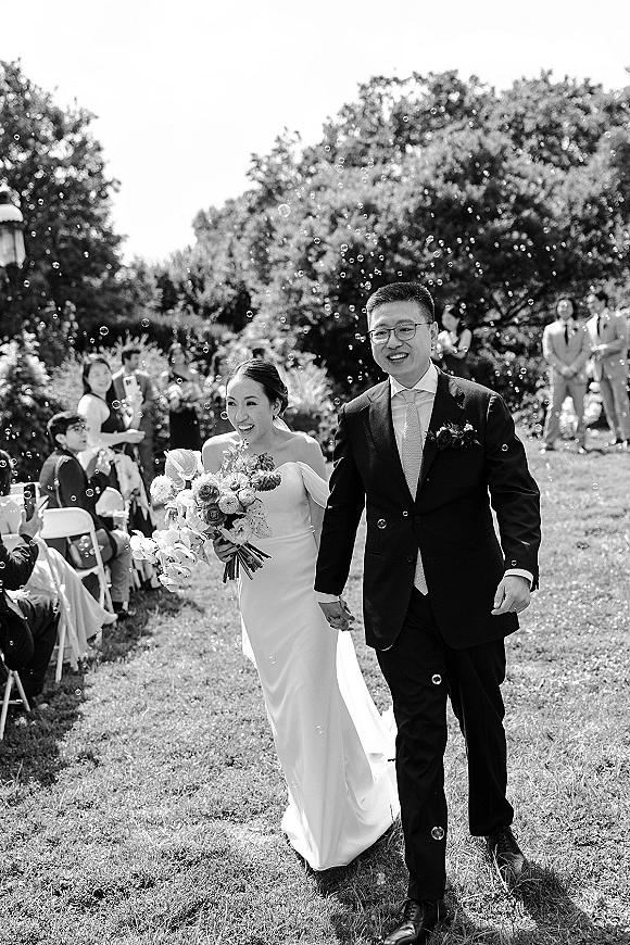 Wedding recessional as bride and groom walk the aisle hand in hand through bubbles, bouquet in hand, guests seated on lawn under trees