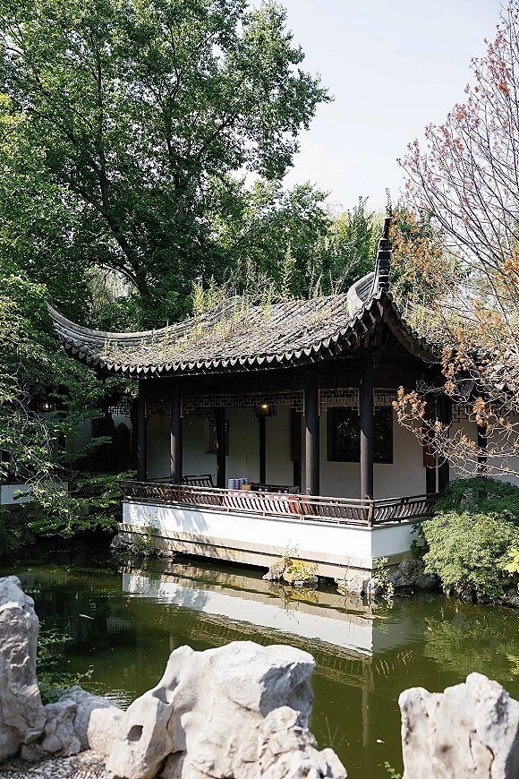 Garden pavilion with traditional pavilion architecture, curved tiled roof and hanging lanterns, set above a pond with rocks and trees