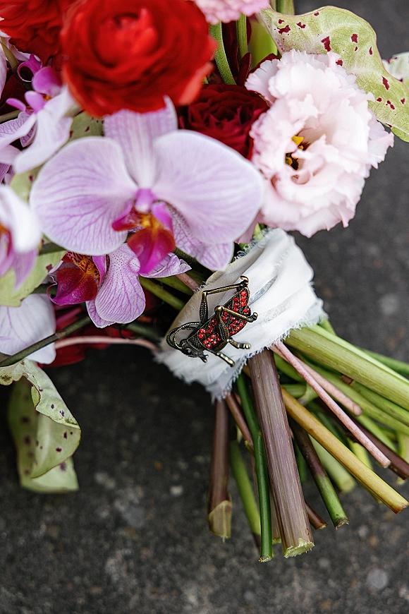Wedding bouquet with orchid blooms, red flowers, greenery and a jeweled brooch on the ribbon wrap resting on dark pavement