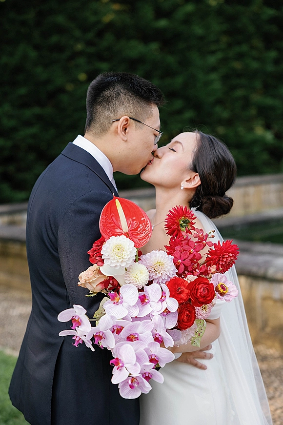 Wedding kiss portrait of bride and groom kissing in side profile, bride holding a red anthurium bouquet, groom in glasses by green hedges