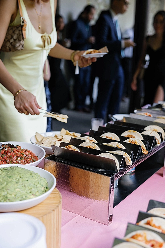 Wedding taco bar with mini tacos in holders, guacamole and salsa bowls, chips, and tongs on a pink tablecloth at an indoor reception