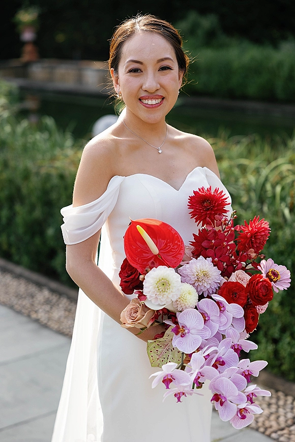 Bridal portrait of a bride holding bouquet in a strapless off-the-shoulder gown with orchid cascade, by garden pond and stone wall walkway
