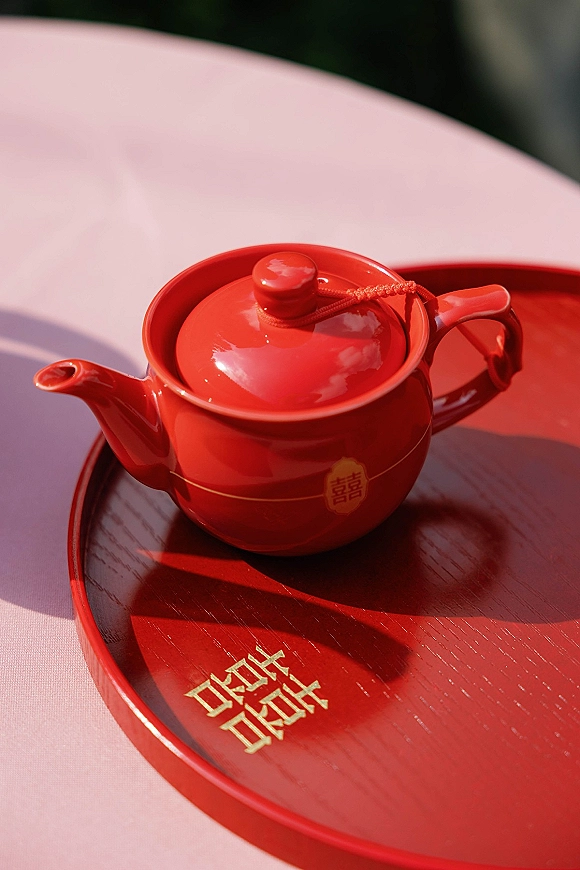 Wedding tea set with a red teapot and gold double happiness symbol on a red serving tray atop a pink tablecloth in sunlight shadows