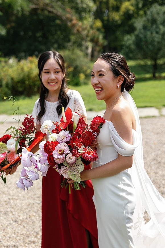 Bride with bridesmaid portrait featuring the bride laughing with bouquets, veil and off-shoulder gown beside a red-dressed bridesmaid on a garden path