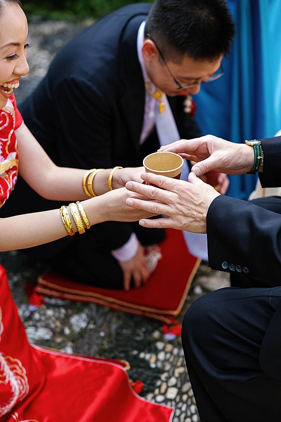 Tea ceremony with bride in red cheongsam serving tea to kneeling groom on a red cushion in an outdoor stone courtyard setting