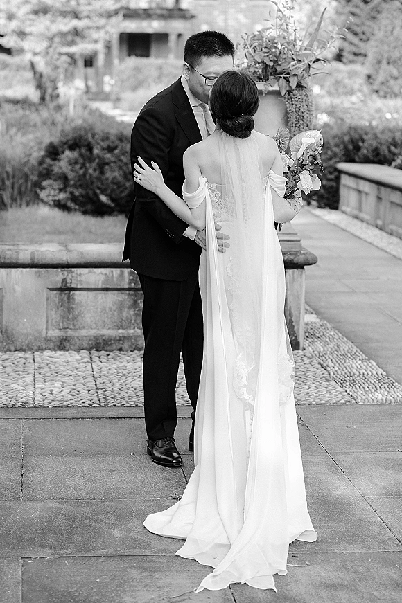 Wedding kiss portrait of bride and groom embracing as she holds a calla lily bouquet, long veil flowing on a stone terrace in a garden courtyard
