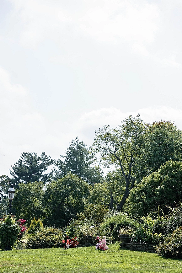 Garden wedding ceremony with floral arrangements and scattered petals lining a grassy aisle, framed by urn planters and trees