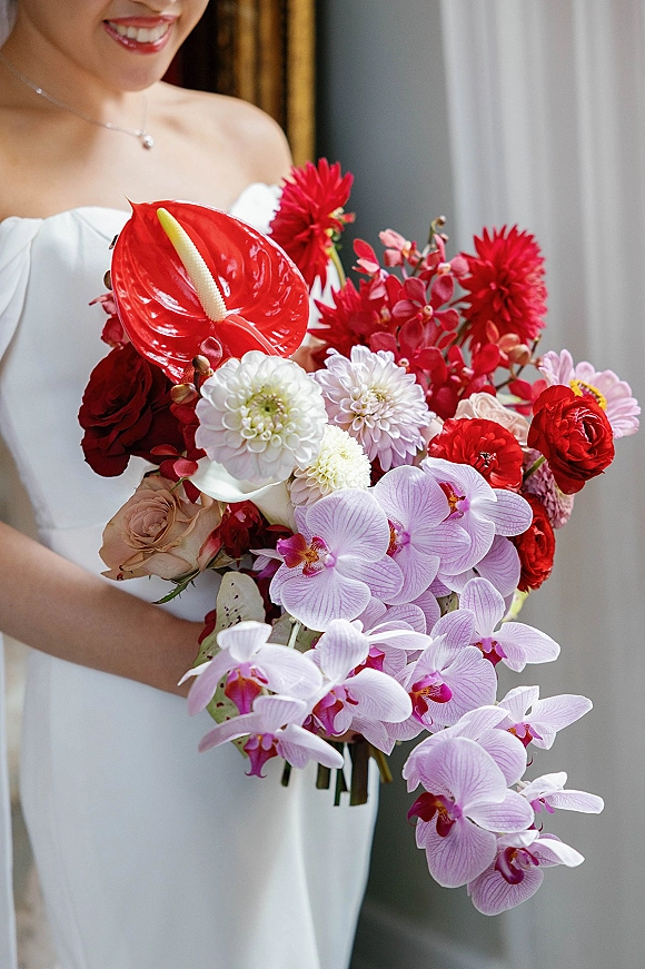 Bridal bouquet with cascading orchid blooms and red dahlias, tied with ribbon stems, held by a bride in soft window-lit indoor light