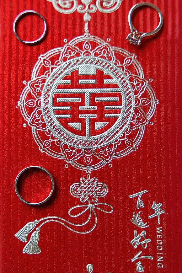 Wedding rings and engagement ring and band resting on red textured fabric with an embossed double happiness symbol and gold foil lettering