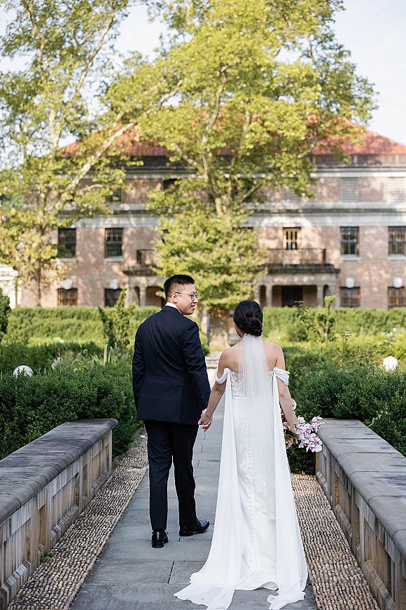 Couple portrait of bride and groom walking away holding hands, bride in long veil with orchid bouquet on a garden walkway by a stone bridge