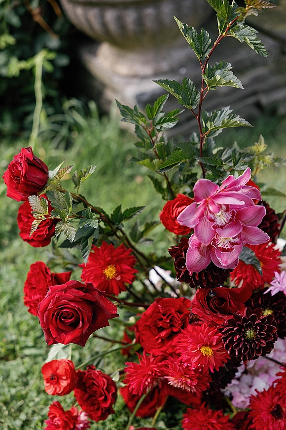 Wedding bouquet with red rose bridal bouquet blooms, dahlias, a pink orchid accent, and lush greenery on a garden lawn near a stone urn planter