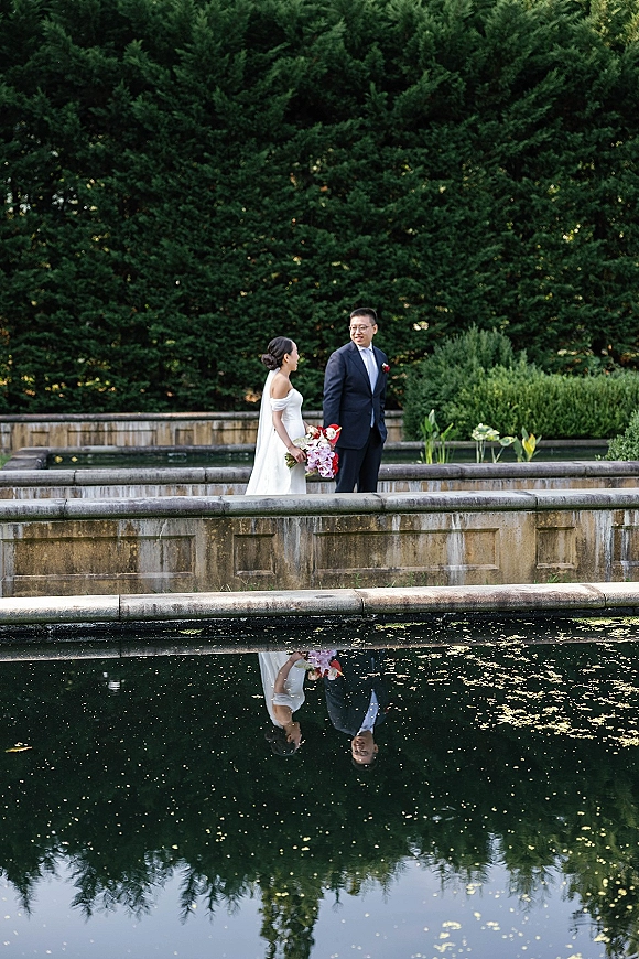 Couple portrait of bride and groom walking by a reflecting pool, bride holding a colorful bouquet with veil, groom in suit by stone balustrade