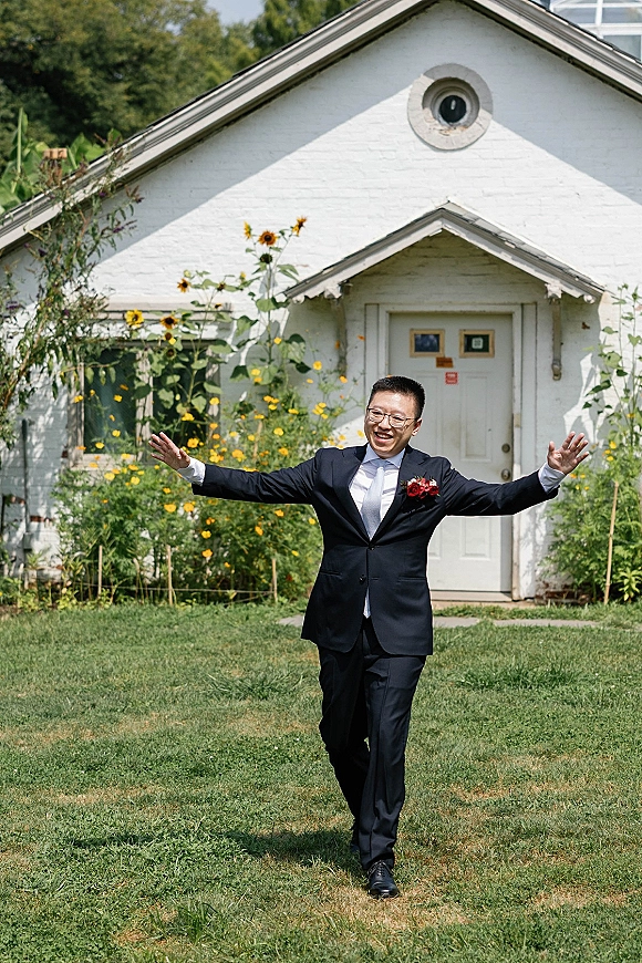 Groom portrait in a navy suit with white tie and boutonniere, smiling with arms outstretched on a lawn by a white chapel.