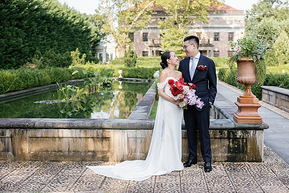 Couple portrait of bride and groom looking at each other, bride holding a red and pink bouquet on a stone terrace by a reflecting pool