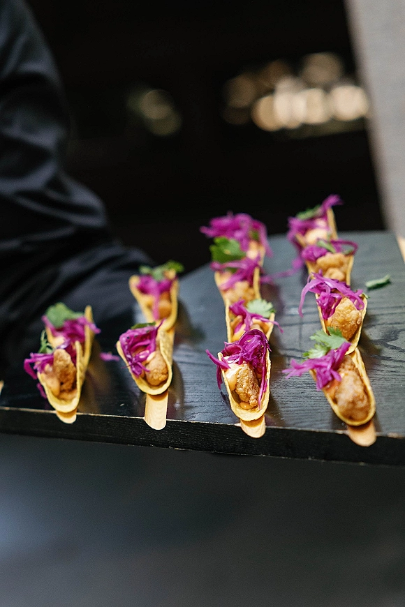Wedding appetizers served as passed hors d'oeuvres, bite-size tacos topped with purple cabbage slaw and cilantro on a wooden tray against dark blur