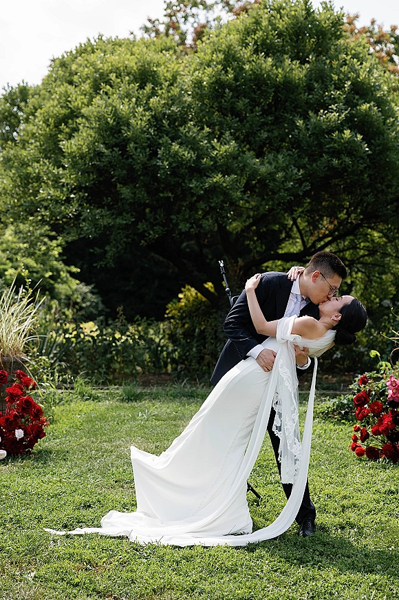 Wedding kiss portrait of bride and groom in a romantic dip, lace veil and red rose florals on a garden lawn by a large tree