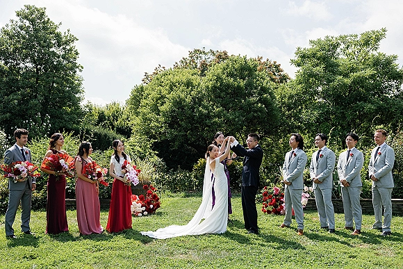 Outdoor wedding ceremony with bride and groom holding hands as bridesmaids in red dresses and groomsmen in grey suits line a garden aisle