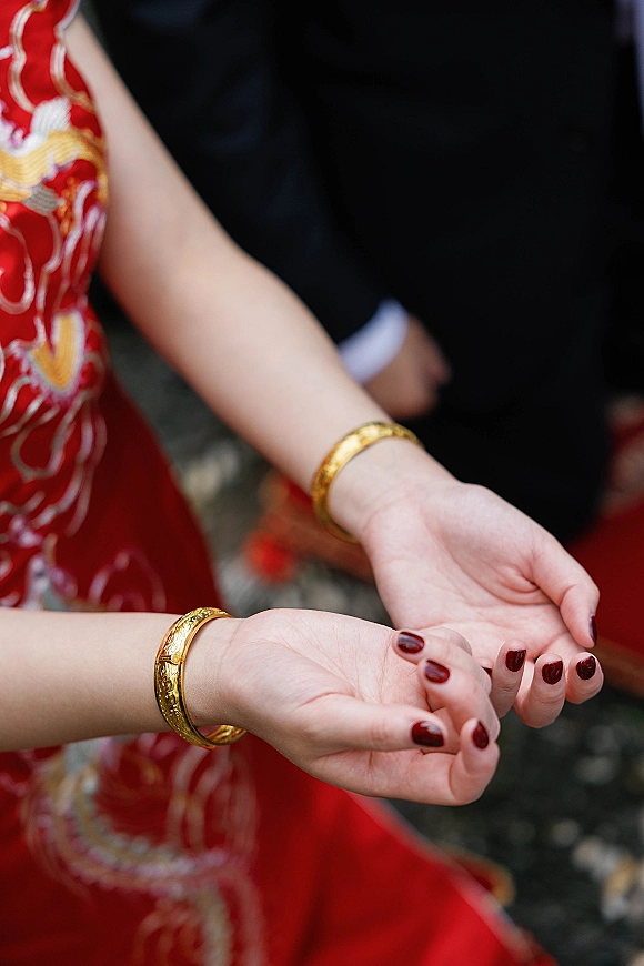 Bridal bangles and a gold bangle bracelet stack on the bride’s wrist, paired with a red embroidered dress and dark red nails outdoors