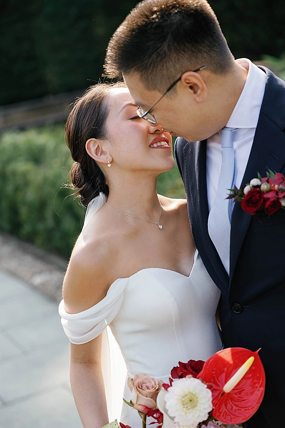 Wedding couple portrait of bride and groom close up touching noses, her veil and bouquet visible as he wears glasses on a garden walkway