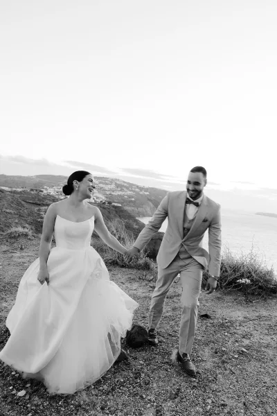 Couple portrait of a bride and groom holding hands on coastal cliffs above the ocean, his bow tie crisp beneath the sky.
