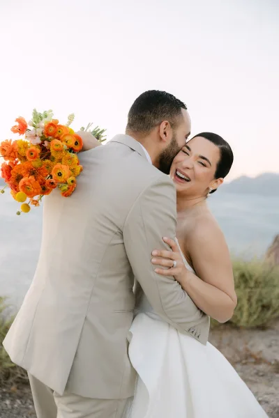 Couple portrait as a bride and groom embrace by a serene waterfront with distant mountains, her orange bouquet vivid between their smiles.