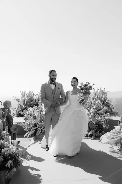 Wedding recessional with a couple walking down the aisle as guests cheer, the bride carrying a cascading bouquet beneath an open sky.