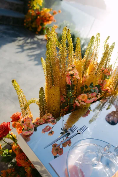 Reception tablescape on a mirrored wedding table with peach and yellow florals, set on a concrete patio with a water view.