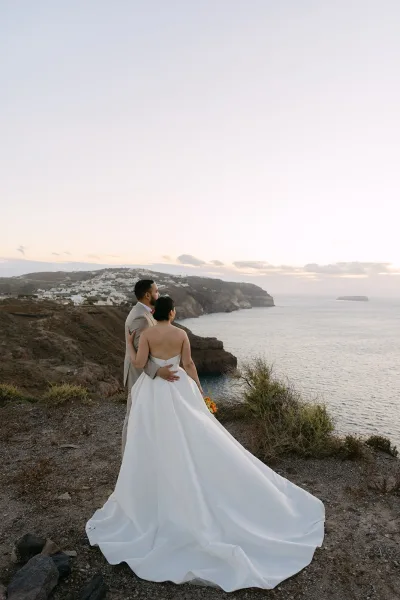 Couple portrait of the bride and groom embracing on coastal cliffs, her orange bouquet vivid against the ocean and distant hillside homes.