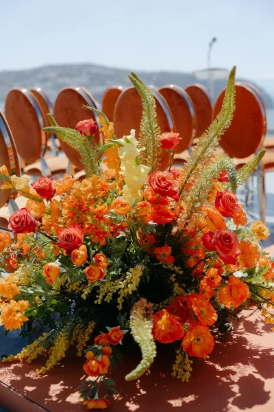 Ceremony flowers in vibrant orange wedding florals cluster around ceremony chairs with lush roses, set against sky, distant hills, and water.