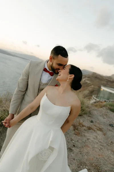 Couple portrait of bride and groom holding hands, touching foreheads, by the ocean cliffs at golden hour, the groom in a sharp red bow tie.