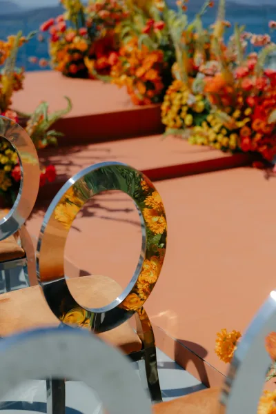 Ceremony seating with modern wedding chairs lines a bright aisle with orange florals, set outdoors against distant hills and open sky.