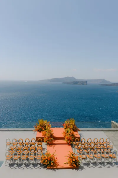 Ceremony setup for an oceanfront wedding ceremony on a terrace, florals edging the aisle before the open sea, clear sky, and distant islands.