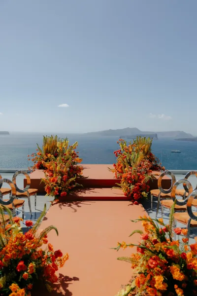 Ceremony setup showcasing oceanfront ceremony decor with orange chair cushions on a terrace overlooking a blue ocean and distant islands.