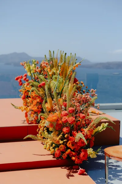 Ceremony flowers anchor outdoor ceremony decor, lush roses on a terrace with glass railing, mountains and an expansive ocean view.