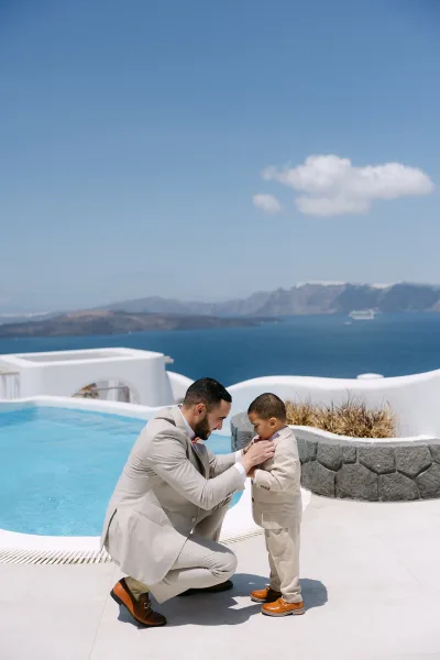 Ring bearer moment with a ring bearer getting ready, adjusting his bow tie beside a pool with an ocean view and white stucco walls under blue sky.