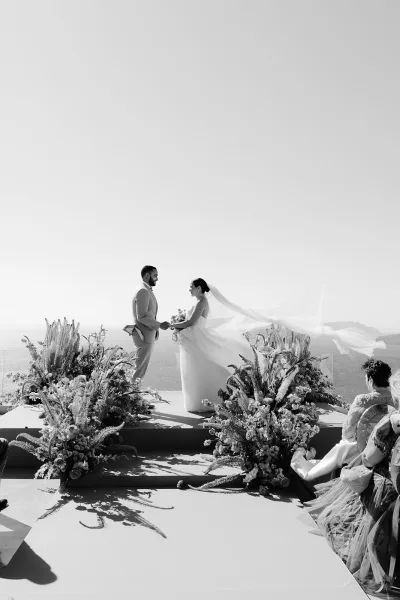 Ceremony moment with wedding vows as the couple stands face to face, her bridal veil catching the breeze, backed by ocean and distant hills.