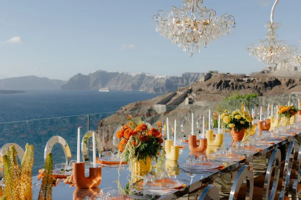 Reception tablescape styled for outdoor reception decor with taper candles, set against a sea view and rocky cliffs beyond a glass railing.