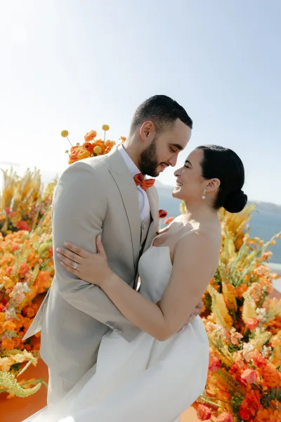 Couple portrait of a bride and groom embrace by the ocean under a blue sky, framed by colorful florals.