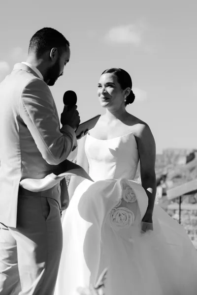 Wedding vows in a tender ceremony moment as the groom speaks into a microphone while the bride listens against a rocky landscape.