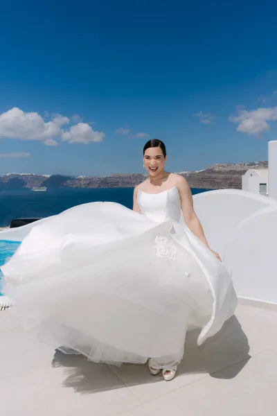 Bridal portrait of a bride twirling her dress on a white terrace above an infinity pool, tulle skirt swirling, ocean and cliffs under a blue sky.