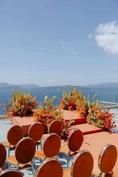 Ceremony setup with outdoor ceremony decor and lush floral arrangements on a terrace, framed by an ocean view, blue sky, and coastal cliffs.