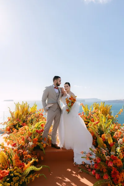 Couple portrait of a bride and groom by the sea, her bold orange rose bouquet bright against a glass-railed terrace and calm coastline.