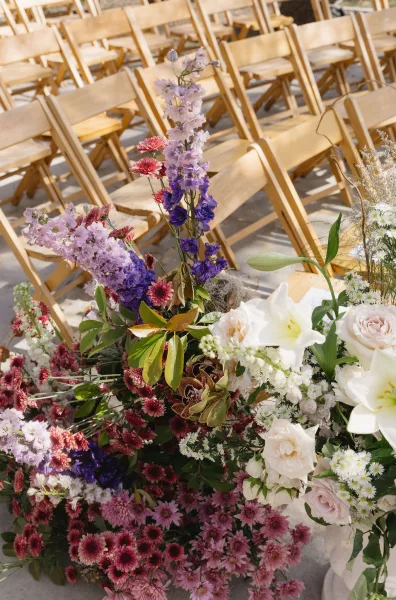 Ceremony aisle flowers shape an elegant aisle floral arrangement with roses, flanking wooden folding chairs on a concrete floor.