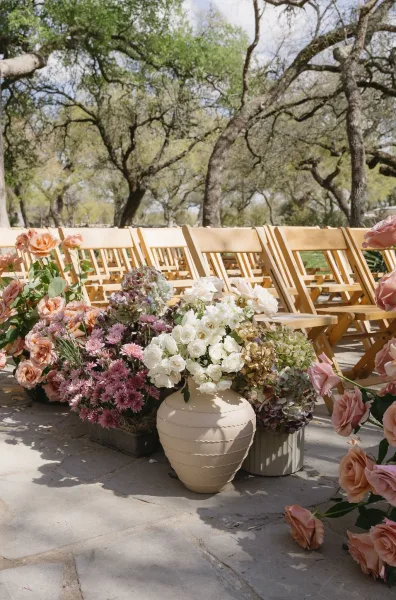 Ceremony aisle decor with artful aisle floral arrangements gathered in a neutral ceramic urn along a stone patio framed by garden trees