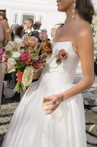 Bridal bouquet, a garden style bridal bouquet with a ribbon wrap, held by the bride as wedding guests gather by a white building facade.