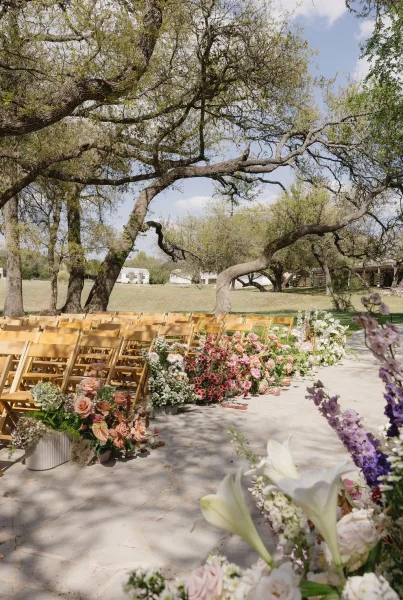 Ceremony aisle flowers frame an outdoor ceremony aisle beside wooden chairs, lush beneath spreading oak trees on a sunlit grass lawn.