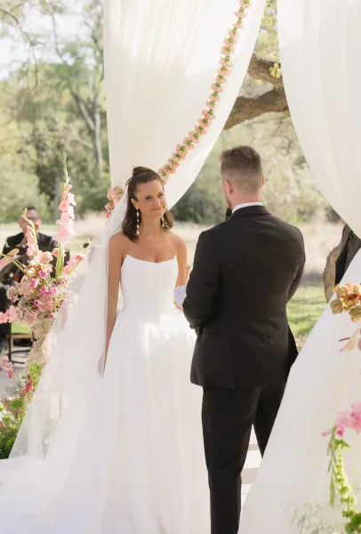 Wedding vows as the bride and groom at the altar share a tender moment beneath a floral arch in a garden, surrounded by trees.