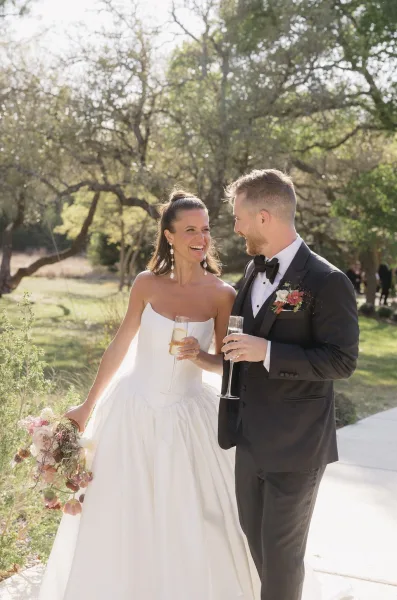 Couple portrait of a bride and groom walking down a garden path beneath trees, laughing, the bride carrying a blush pink bridal bouquet.